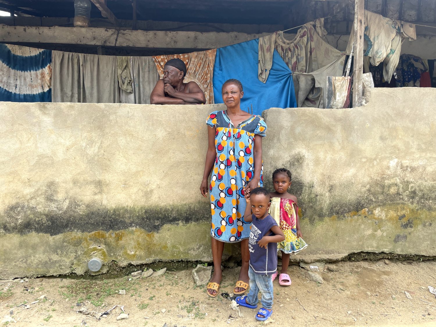 Marie-Claire (ici avec une voisine et ses enfants) a fait construire un mur pour lutter contre l’eau, sans succès. La trace noire indique la hauteur de l’eau lors des dernières inondations © Globe Reporters